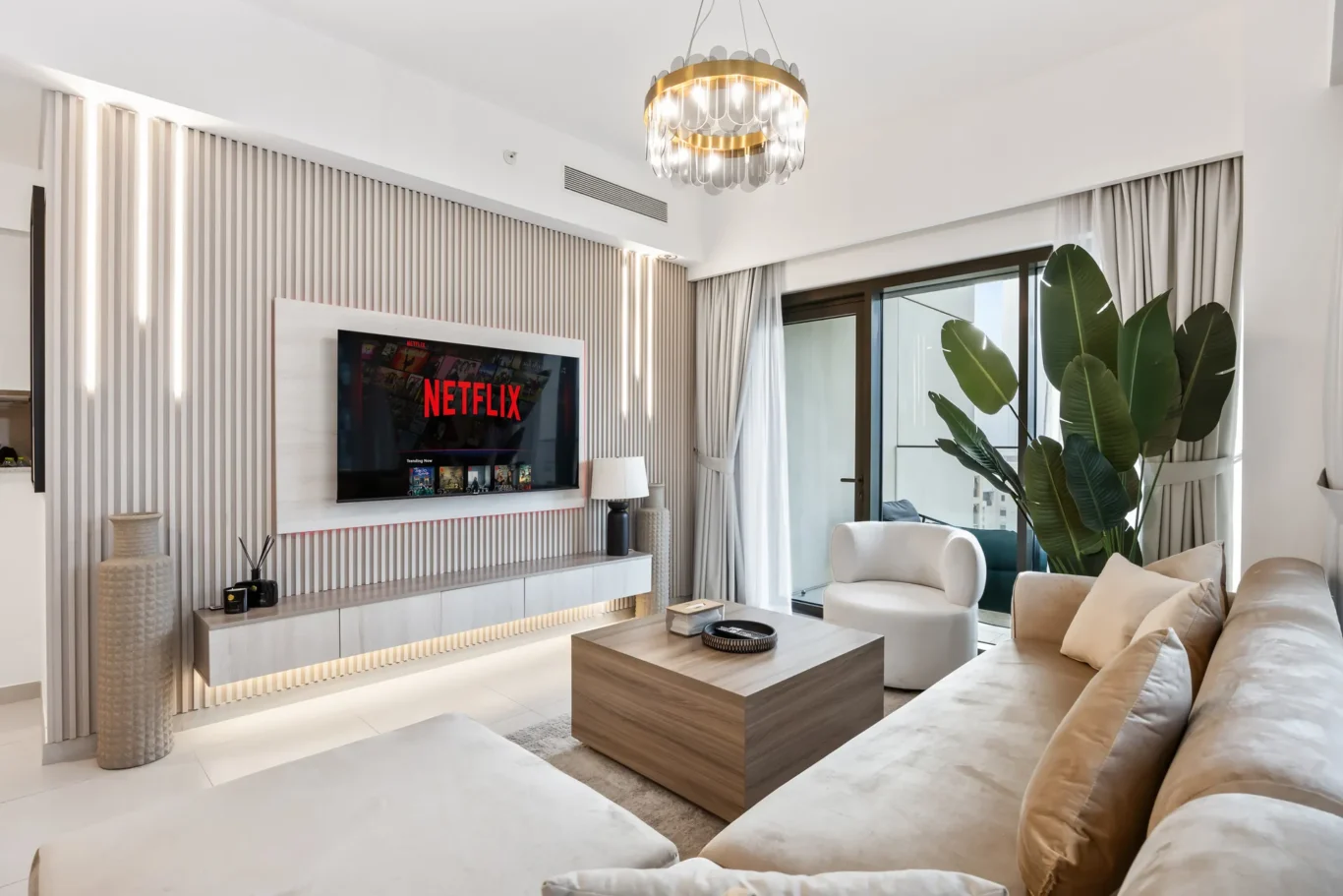Modern living room with a wall-mounted Netflix TV, beige seating, and a tall green plant beside glass doors.