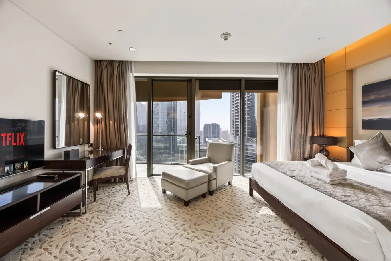 Modern hotel room with a king bed, seating area, and balcony doors revealing a city skyline.