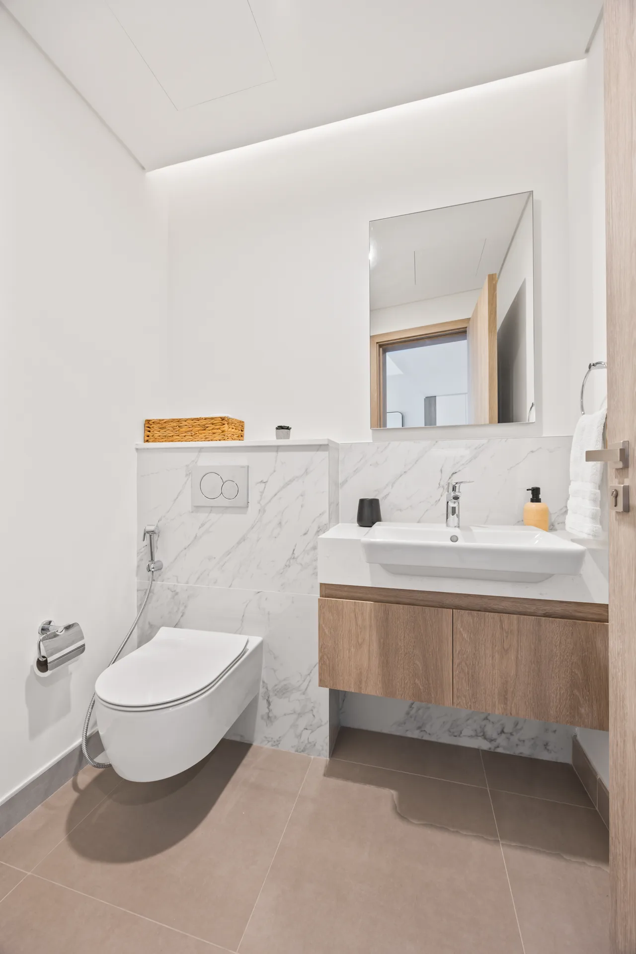 Modern white bathroom with a wall-hung toilet, marble-tiled half-wall, and a wooden vanity under a large mirror.