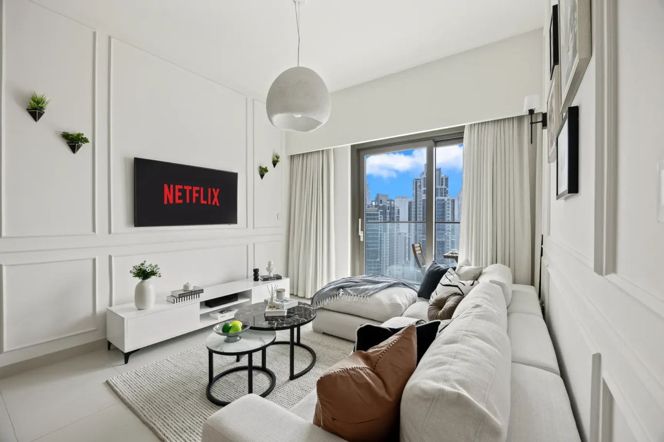 Bright modern living room with white paneled walls, a wall-mounted TV showing Netflix, and a light gray sofa setup next to a balcony with city skyline outside the windows.