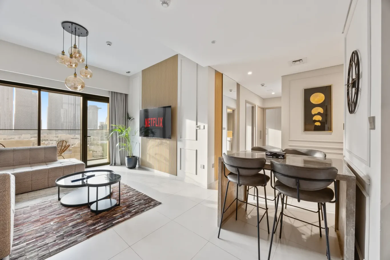 Open-concept living area with a wall-mounted Netflix screen, neutral furniture, and a glass-top coffee table by a patterned rug and large window view.