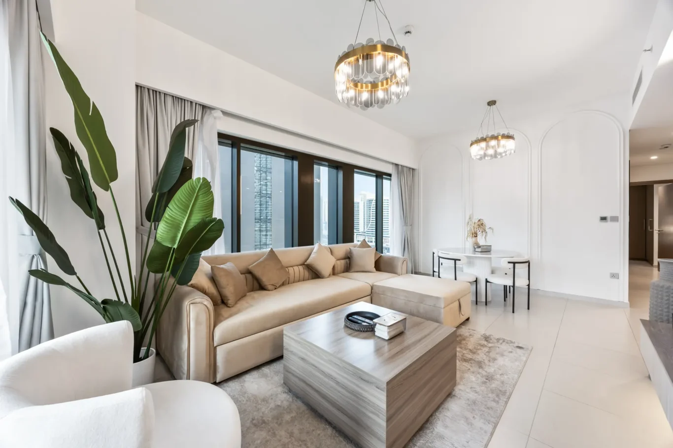Bright, modern living room with a beige sectional sofa, large green plants, and a wood coffee table facing tall windows.