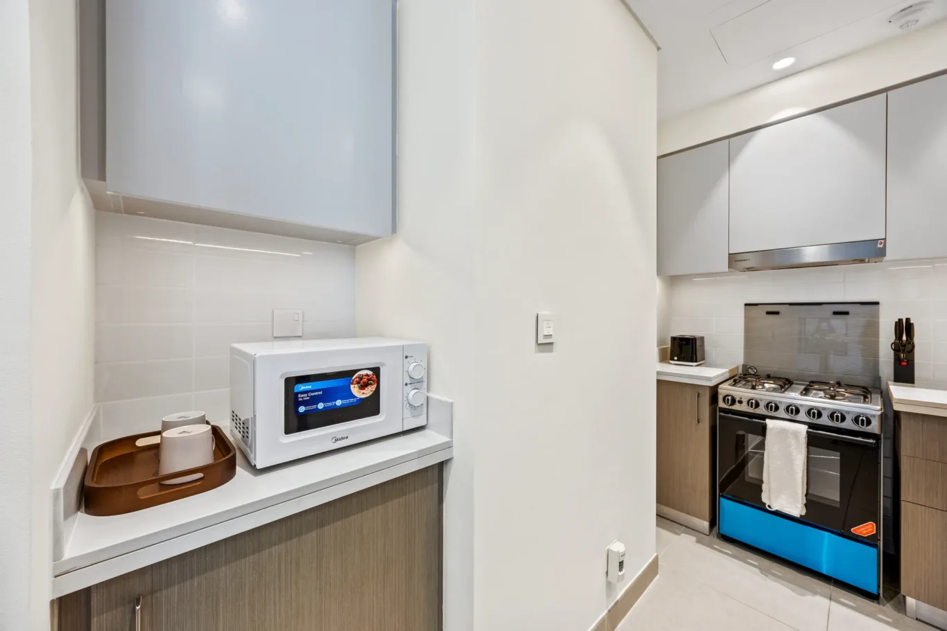Compact kitchen with a white microwave on a counter and a wooden tray nearby, amid light cabinets and tiled backsplash.