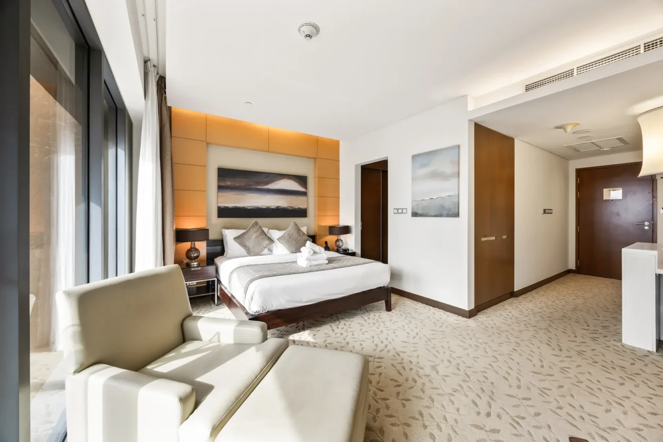Hotel room with a king bed, warm backlit headboard, two nightstands, and a seating area by large windows with white curtains.