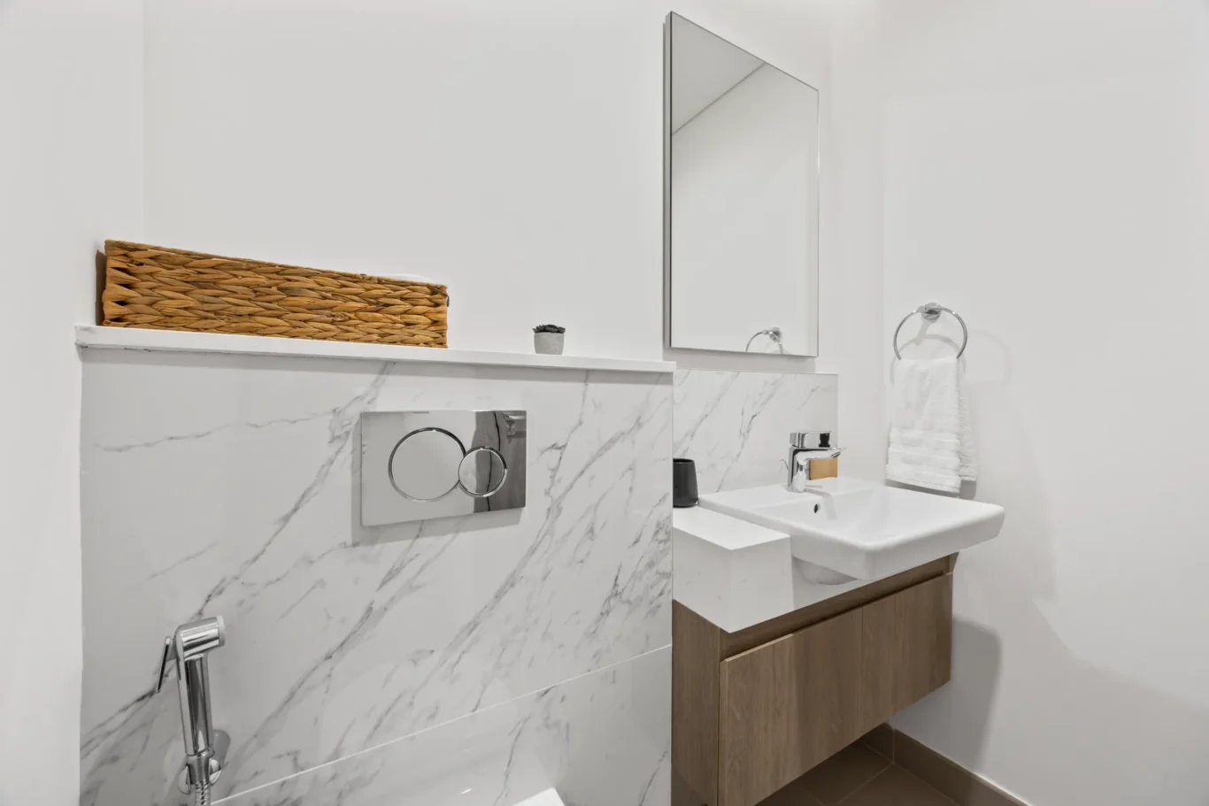 Modern bathroom featuring white marble walls and countertop, a wall-mounted chrome toilet panel, a wooden vanity, and a square mirror above the sink.