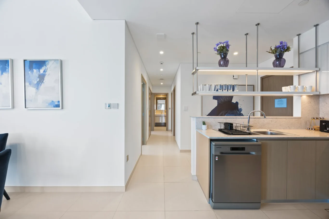 Modern open-plan kitchen with a central island, hanging shelves with vases of purple flowers, and a hallway leading to back rooms