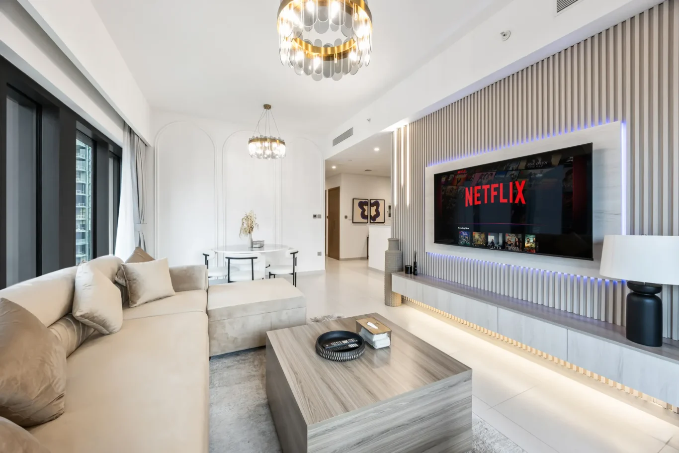 Modern living room with beige sectional sofa, glass windows, and a large wall-mounted TV showing Netflix with blue lighting accents.