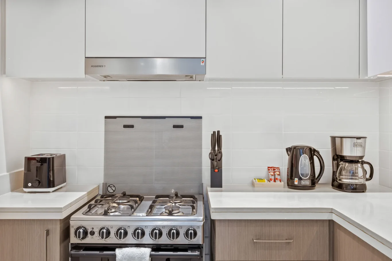 Modern kitchen counter with a stainless gas range, white tile backsplash, and appliances (toaster oven, kettle, and coffee maker) on either side.