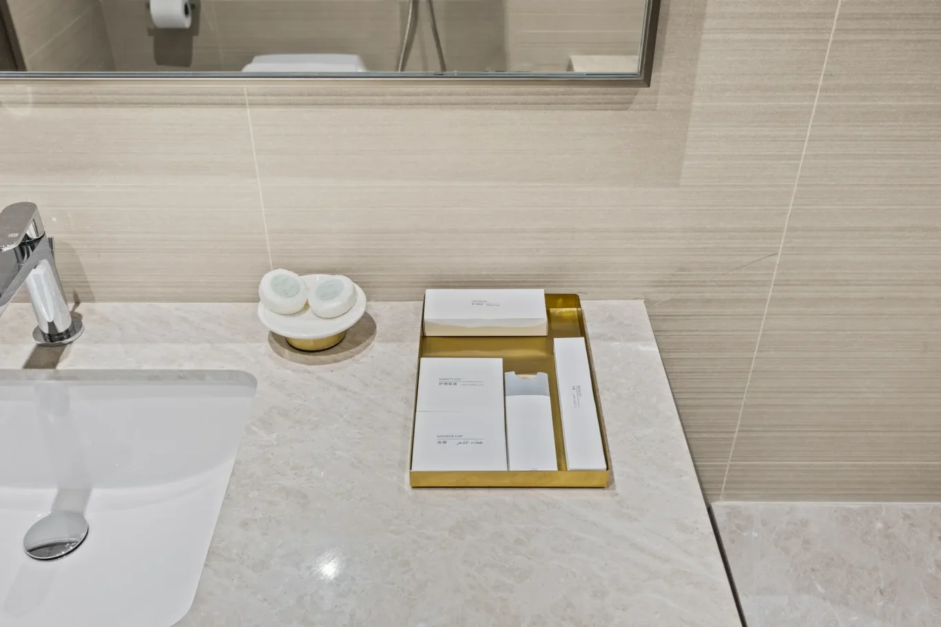 Sleek bathroom vanity with a chrome faucet, marble countertop, and a gold tray holding boxed toiletries beside the sink mirror checks in.