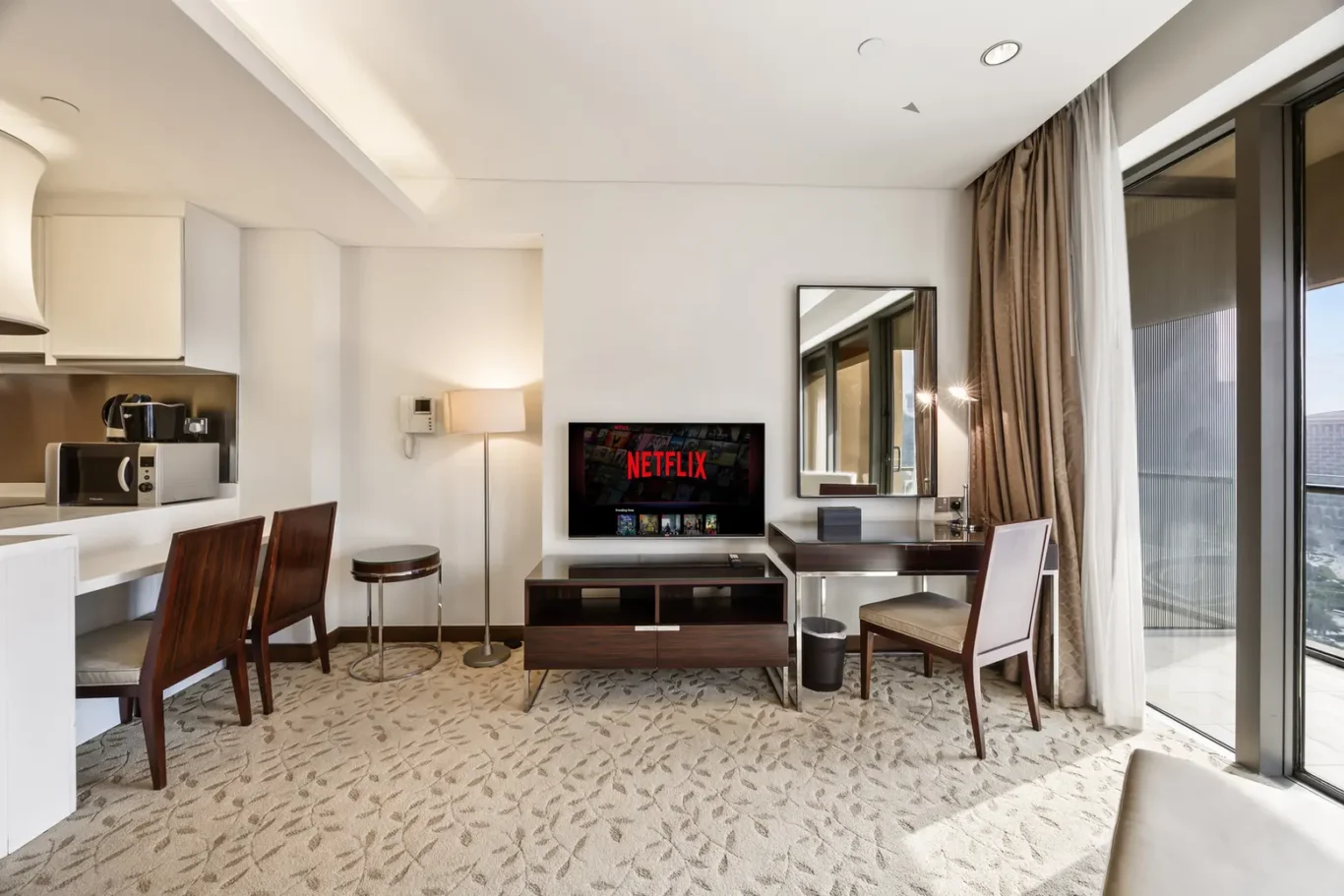 Hotel suite living area with a TV on the wall showing Netflix, a glass-top console, desk with chair, and a balcony window to the right.