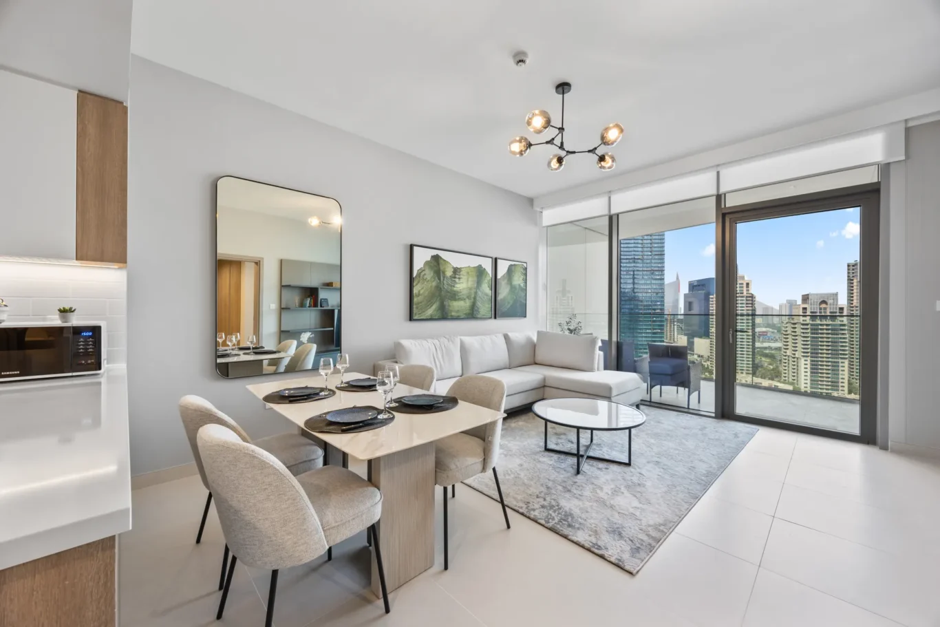 Modern open-plan living and dining area with a marble table, beige chairs, and a gray sofa, overlooking a city skyline through glass balcony doors and windows.