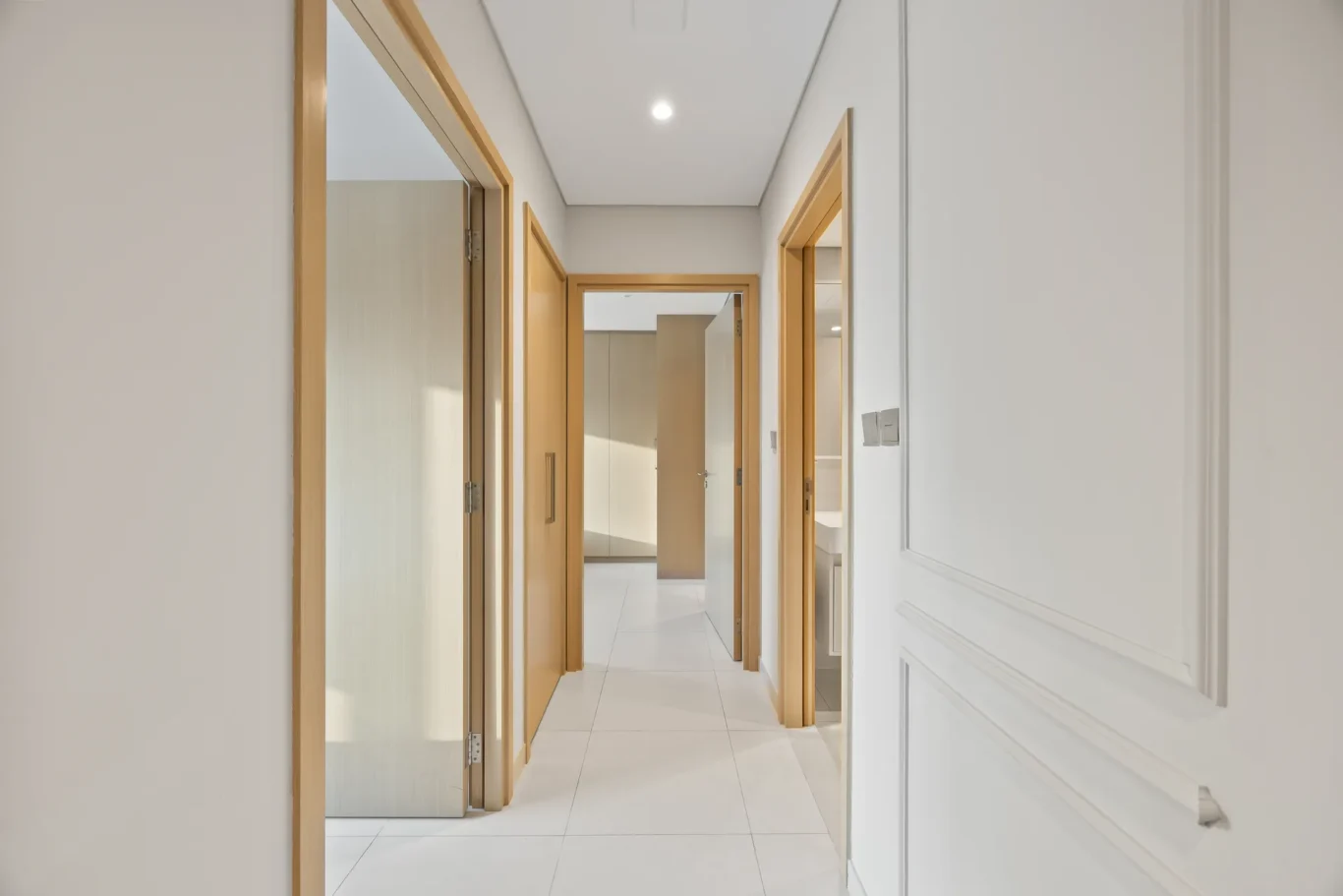 Bright hallway with white walls and light wood door frames, doors open to rooms along a tiled floor.