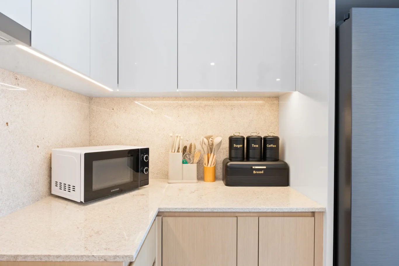 Modern kitchen counter with a white microwave, utensil holder, and labeled canisters for sugar, tea, and coffee nearby a bread toaster.