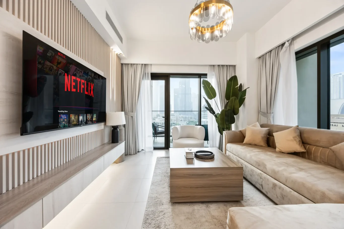 Living room with a wall-mounted TV showing Netflix, beige sectional sofa, and a wooden coffee table by large windows with curtains and a potted plant nearby.