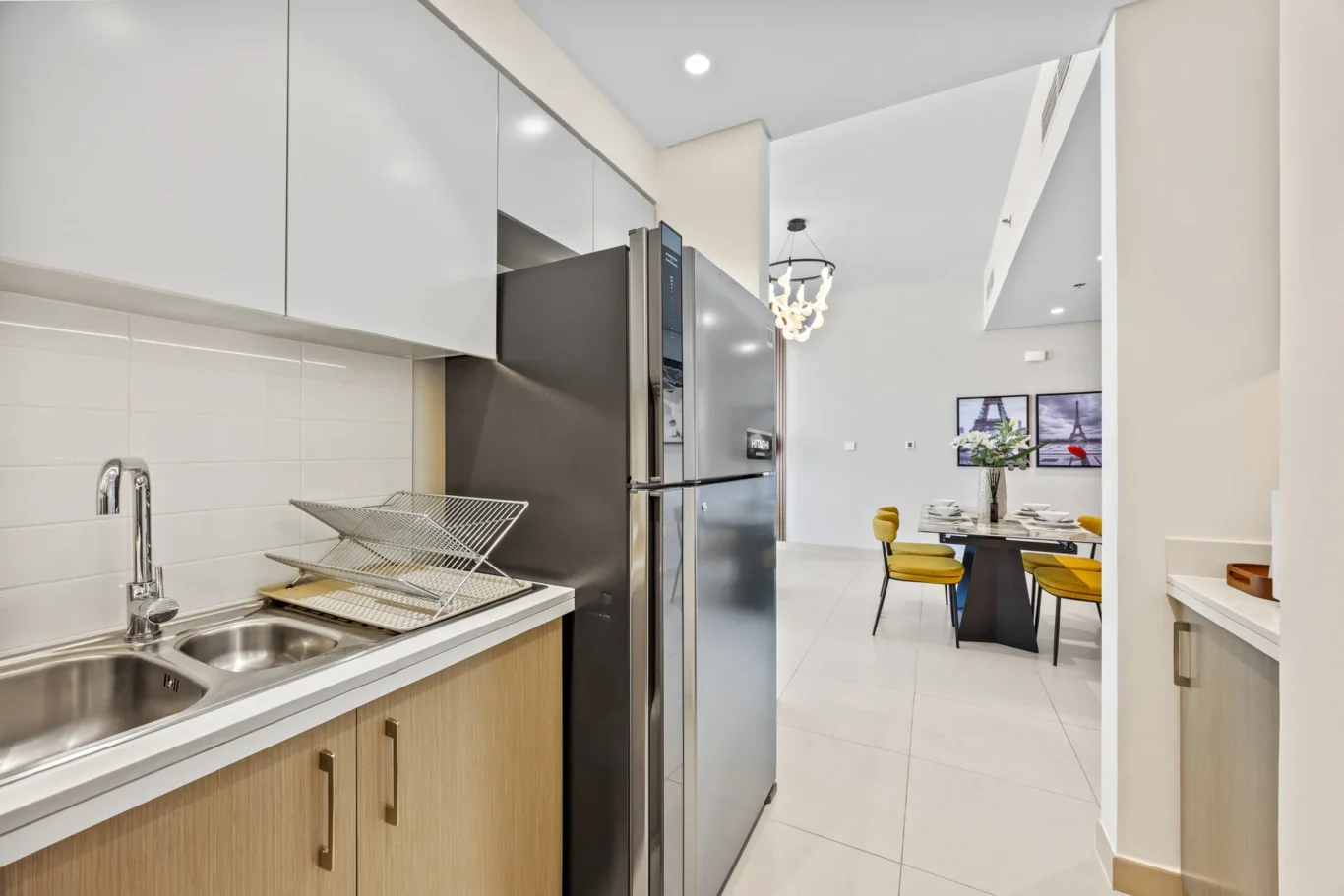 Modern kitchen with a stainless steel fridge, white cabinets, and a dish rack by the sink; dining area visible beyond.