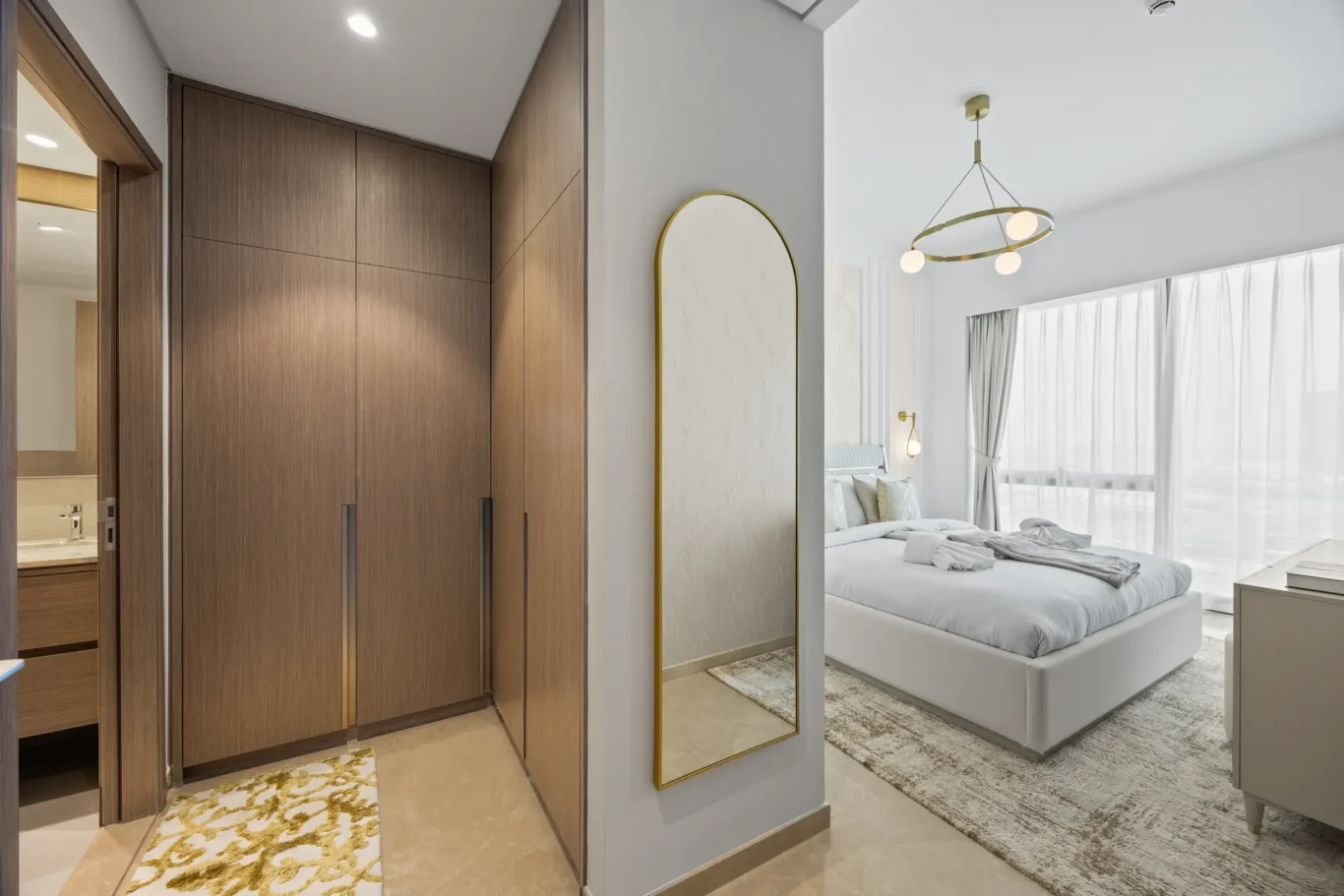 Hallway with tall wooden wardrobes and a gold-framed arched mirror, leading to a bright bedroom with a round gold chandelier and large windows.