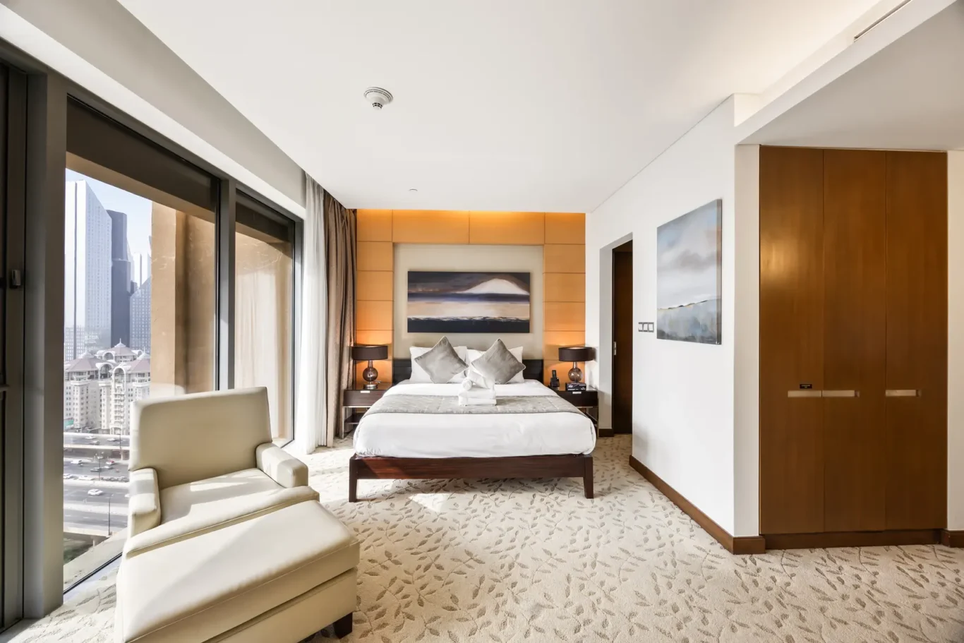 Hotel room with a king bed, orange paneled headboard, two nightstands, and a city skyline view through large windows behind seating area.