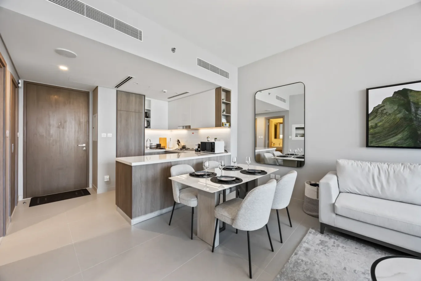 Open-plan living area featuring a kitchen island, six-seat dining table with place settings, and a mirrored wall near a gray sofa across from artwork.