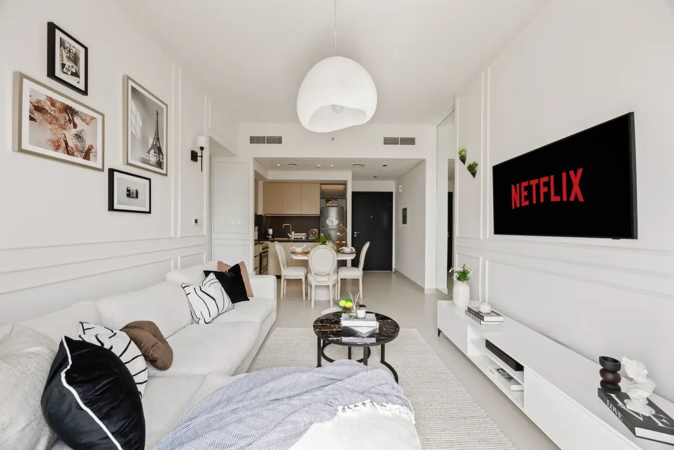 Bright modern living room with white walls, a large sofa with black-and-white cushions, and a wall-mounted Netflix screen.
