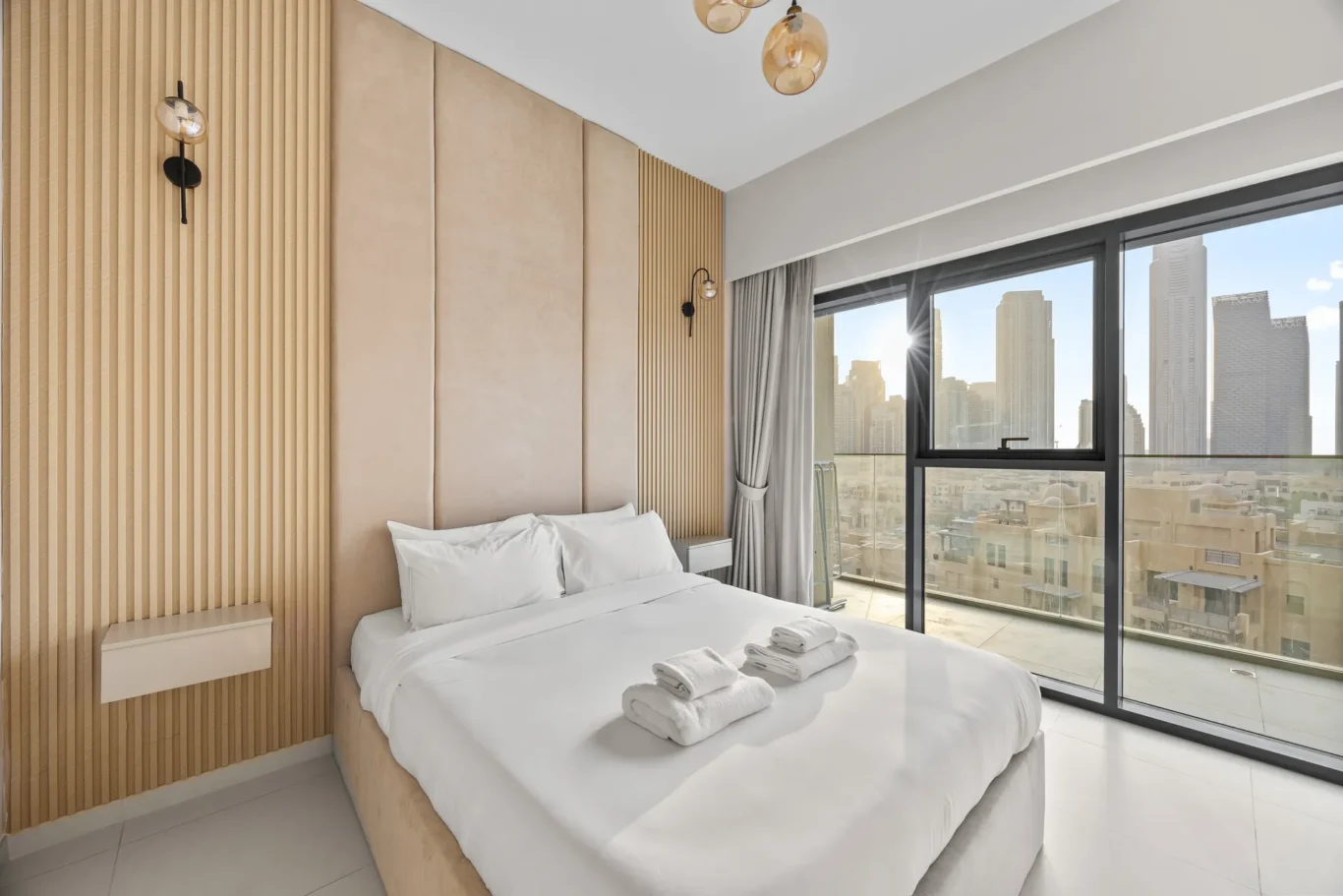 Hotel bedroom with a large window showing a city skyline; white bed, beige padded headboard, and wooden slat wall accents.