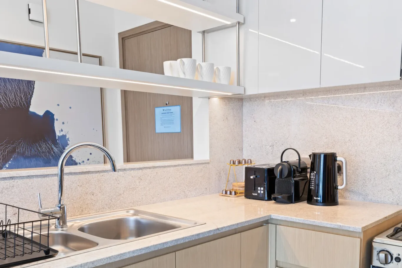 Modern kitchen counter with a double sink, chrome faucet, dish rack, and small black appliance set (toaster, coffee machine, kettle) on a light granite surface.