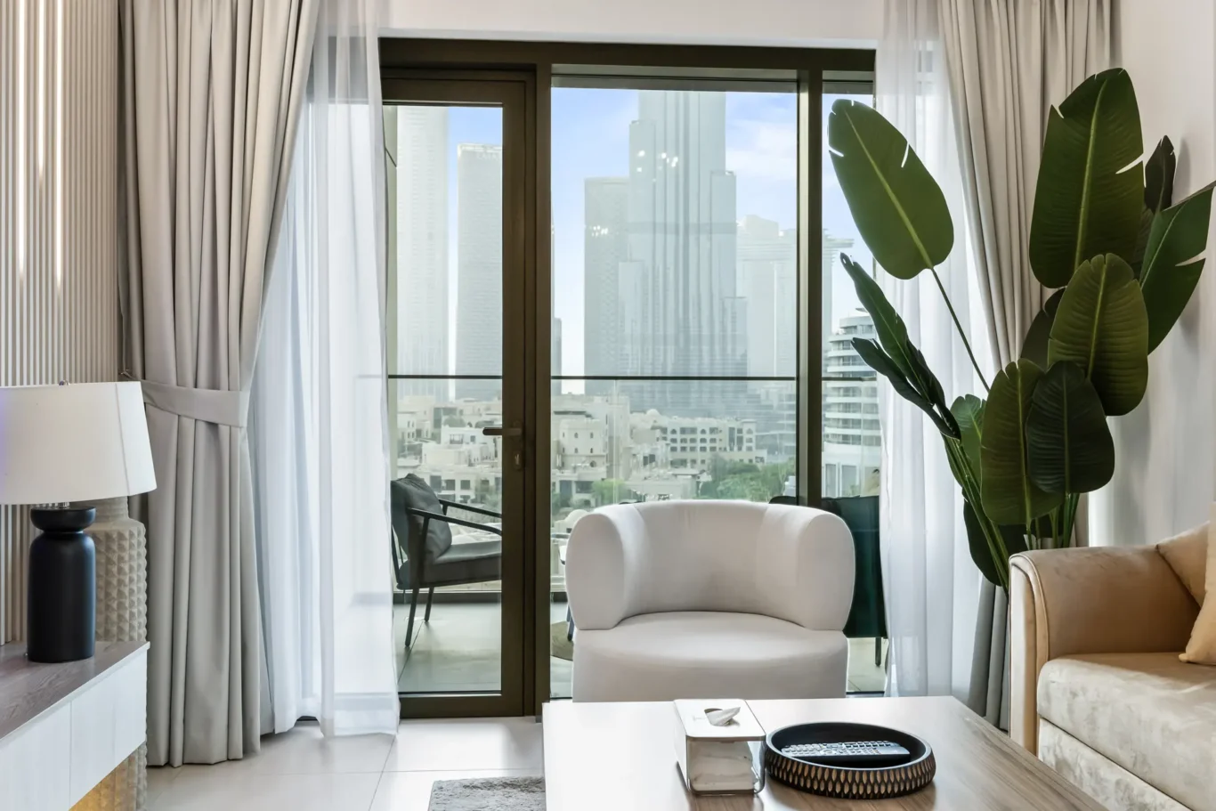 Living room with a glass balcony door opening to a city skyline, white chair, and tall green plant.