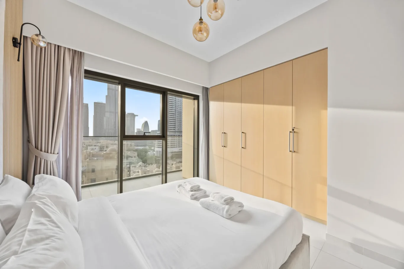Bright hotel bedroom with white bedding, folded towels on the bed, and large windows revealing a city skyline.