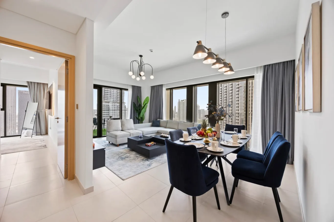 Open-plan living and dining area in a modern apartment with a gray sectional, navy dining chairs, and a pendant-light row; large windows overlook a city skyline.
