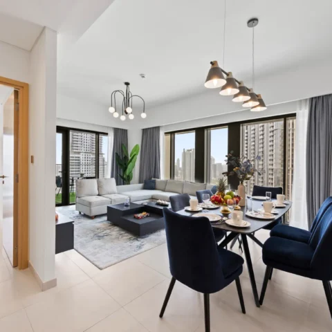 Open-plan living and dining area in a modern apartment with a gray sectional, navy dining chairs, and a pendant-light row; large windows overlook a city skyline.