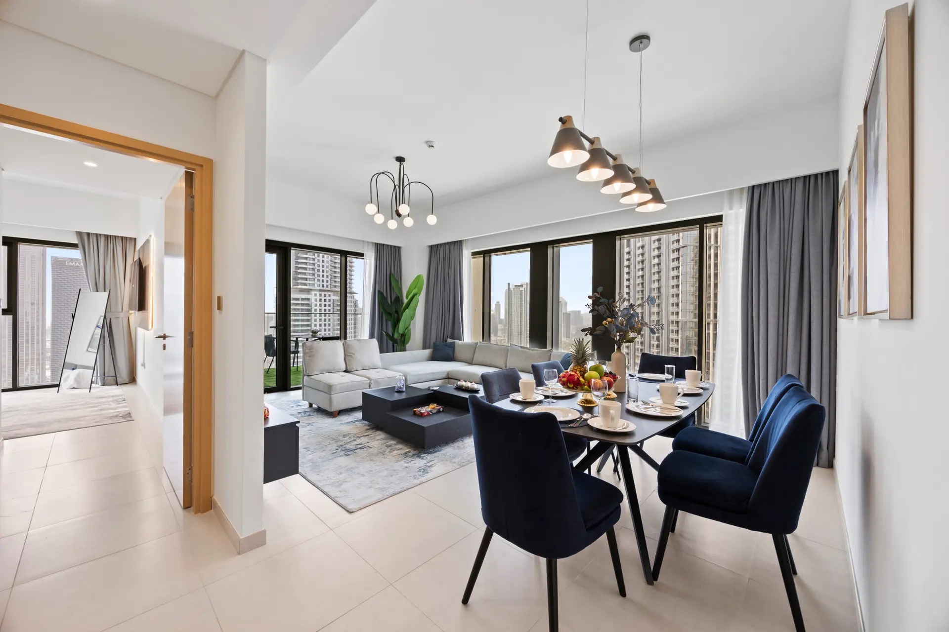 Open-plan living and dining area in a modern apartment with a gray sectional, navy dining chairs, and a pendant-light row; large windows overlook a city skyline.