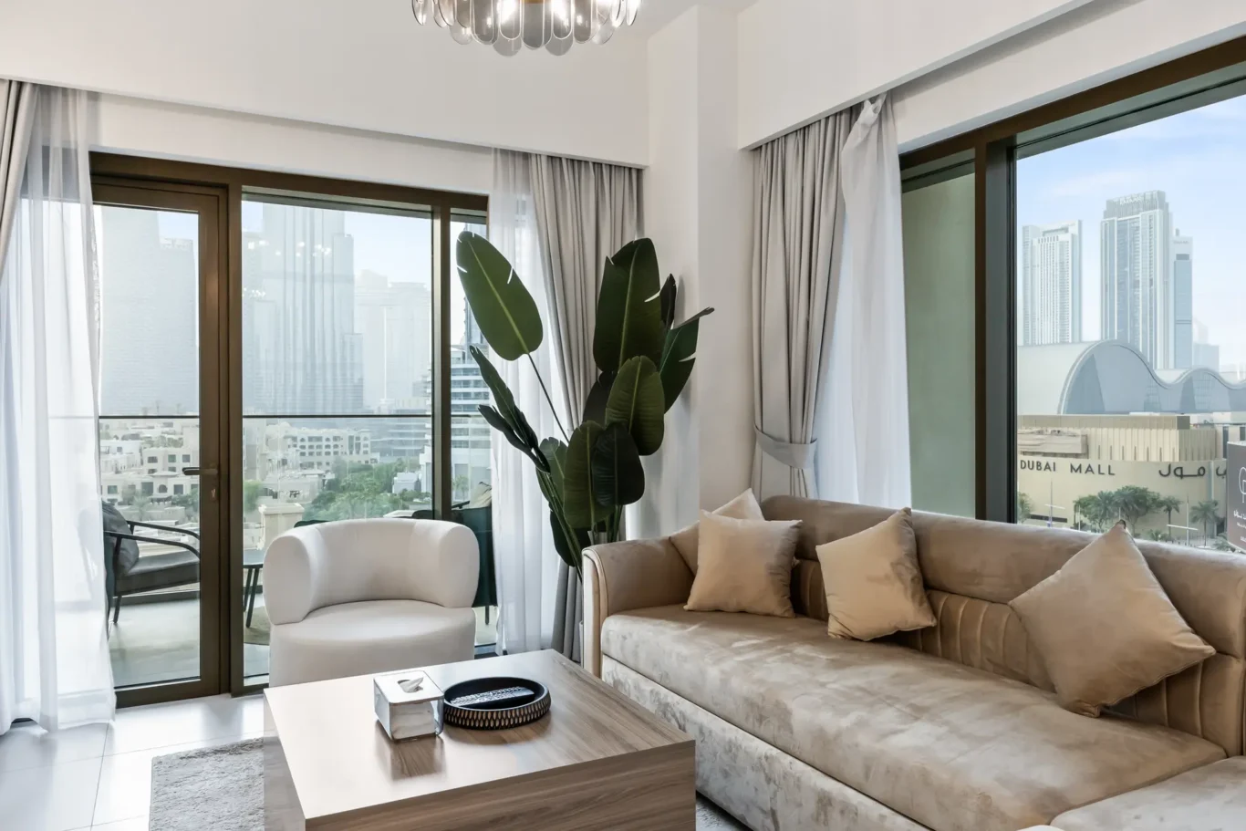Bright living room with a beige sectional sofa, white armchair, and a tall leafy plant by glass doors opening to a balcony with a city view.