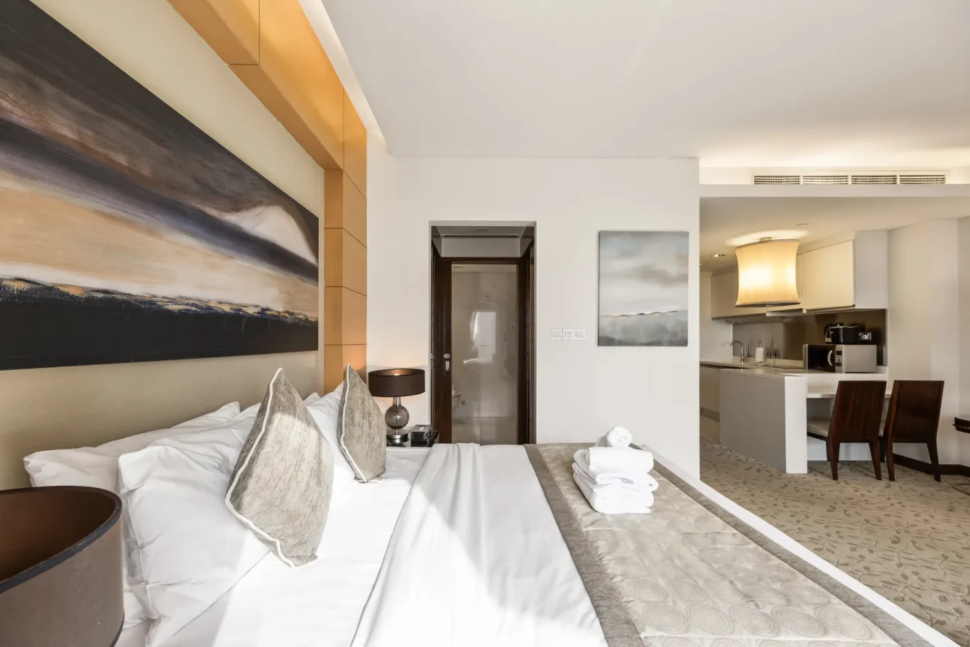 Modern hotel suite: large bed with white linens and gray pillows, nightstand lamp, and an open-plan kitchen/dining area in the background.