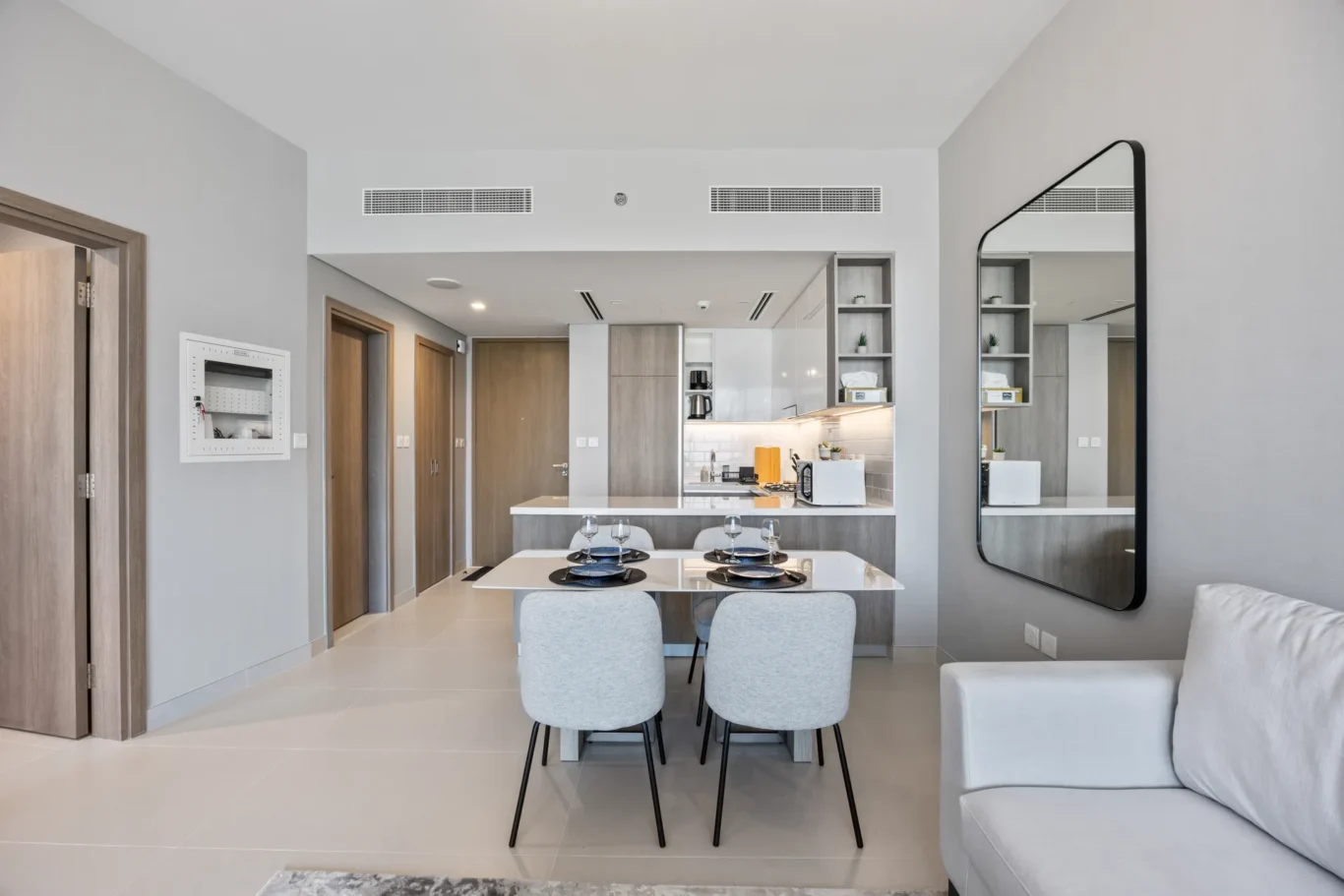 Open-plan dining and kitchen area with a white table set for four, gray upholstered chairs, and a large rounded-frame mirror to the right.