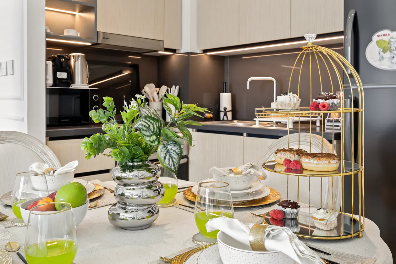 Modern kitchen breakfast table with green drinks, fruit bowl, and a gold three-tier pastry stand holding cupcakes and donuts.
