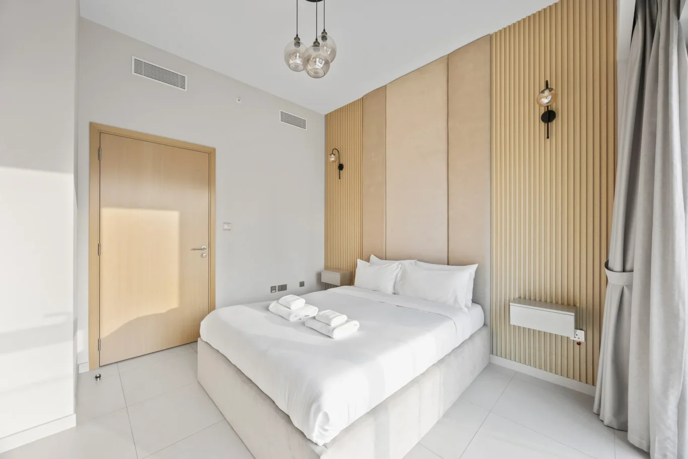 Modern hotel room with white bedding, a low platform bed, beige wood slat headboard, and a wooden door in view of the room.