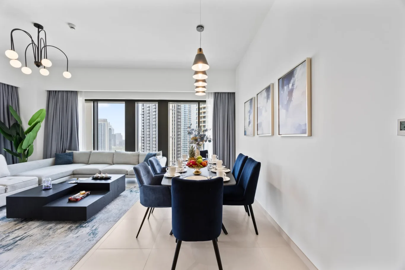 Bright open-plan living and dining area with a city skyline view, modern chandelier, and blue dining chairs nearby.