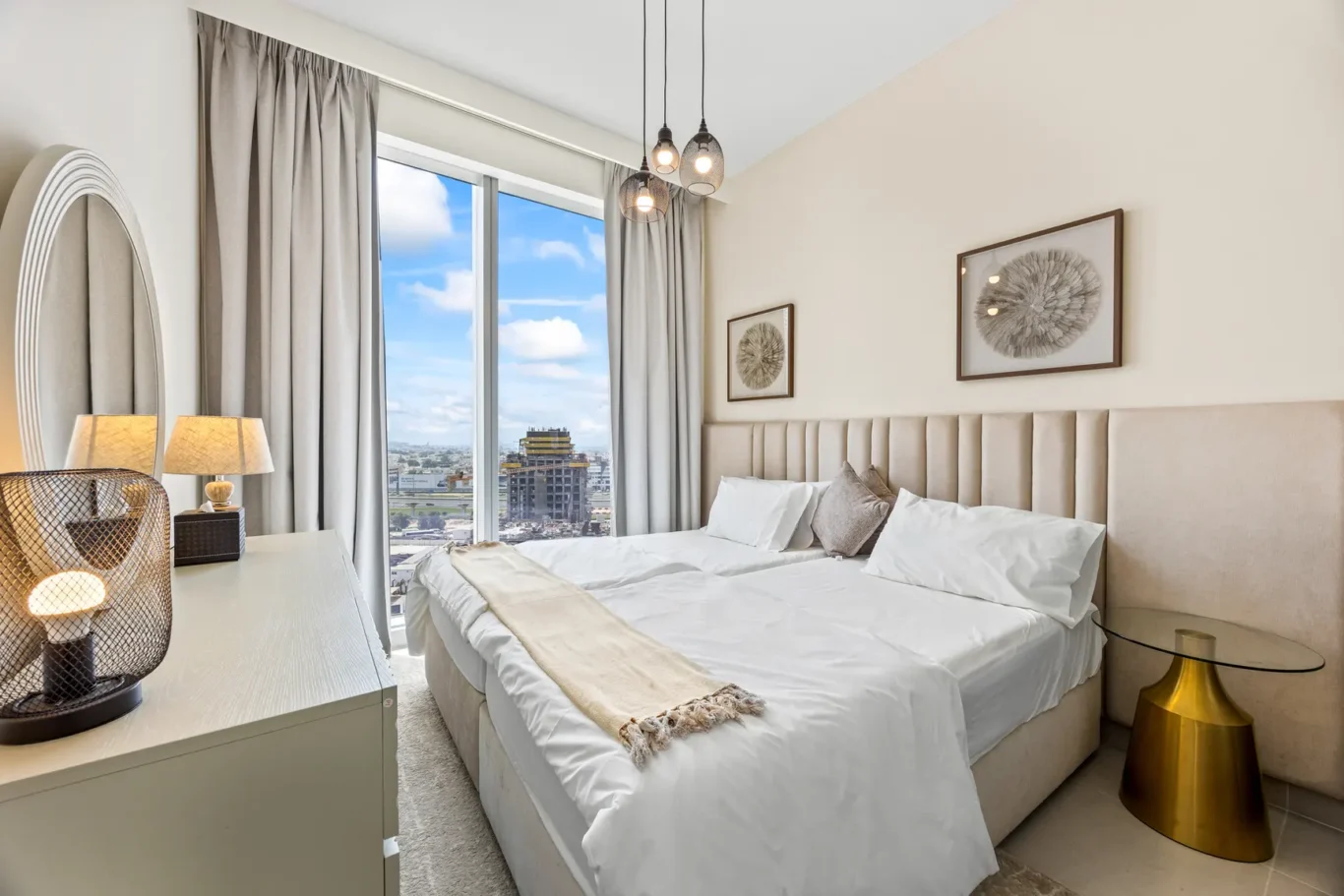 Hotel room with a large window view of the city, beige upholstered headboard, white bedding, and framed circular art pieces above the bed.