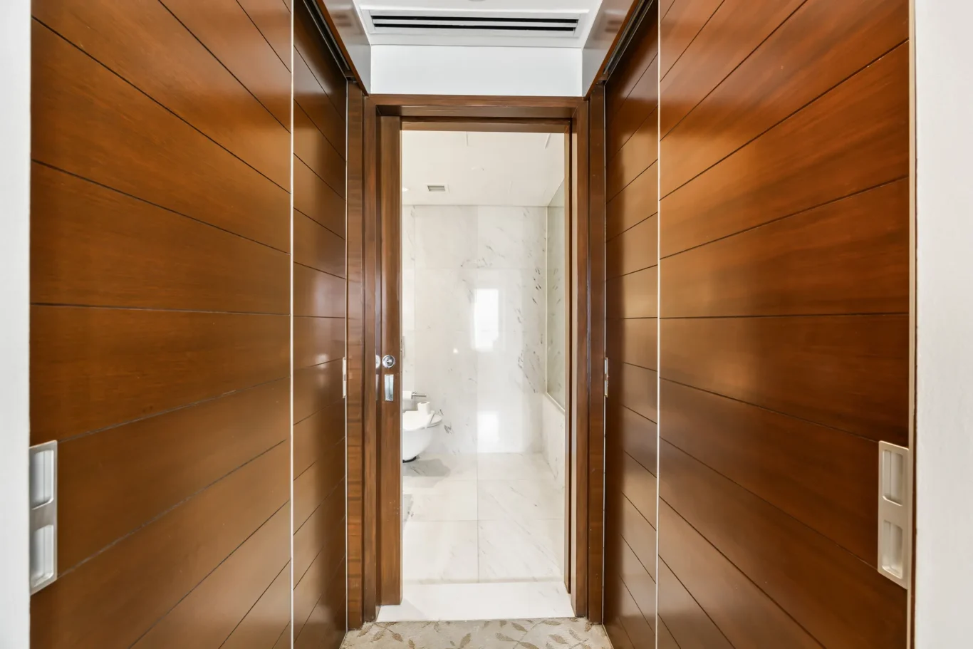 Wood-paneled hallway opening into a marble-tiled bathroom, with a door frame in the center and bathroom fixtures visible beyond.