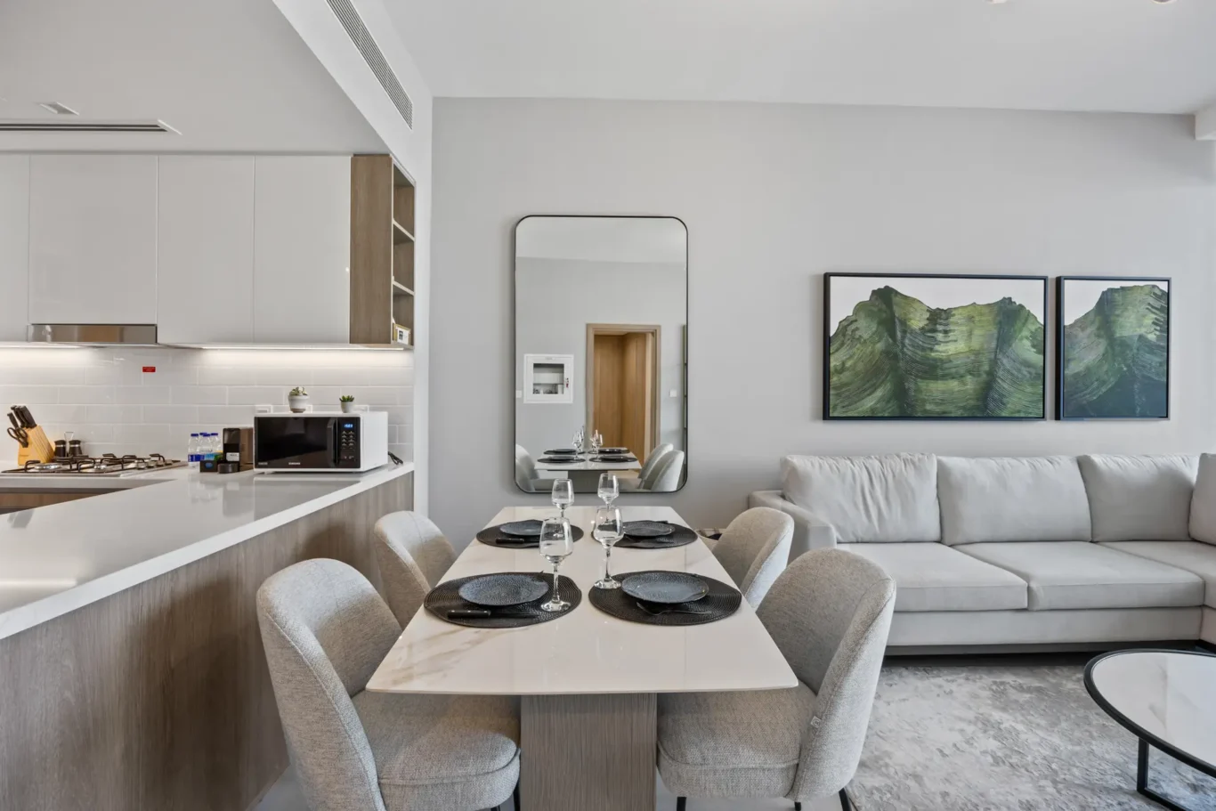 Modern open-concept kitchen and dining space with a marble table set for six, gray upholstered chairs, white cabinets, and a living area with a gray sofa and mountain art on the wall.
