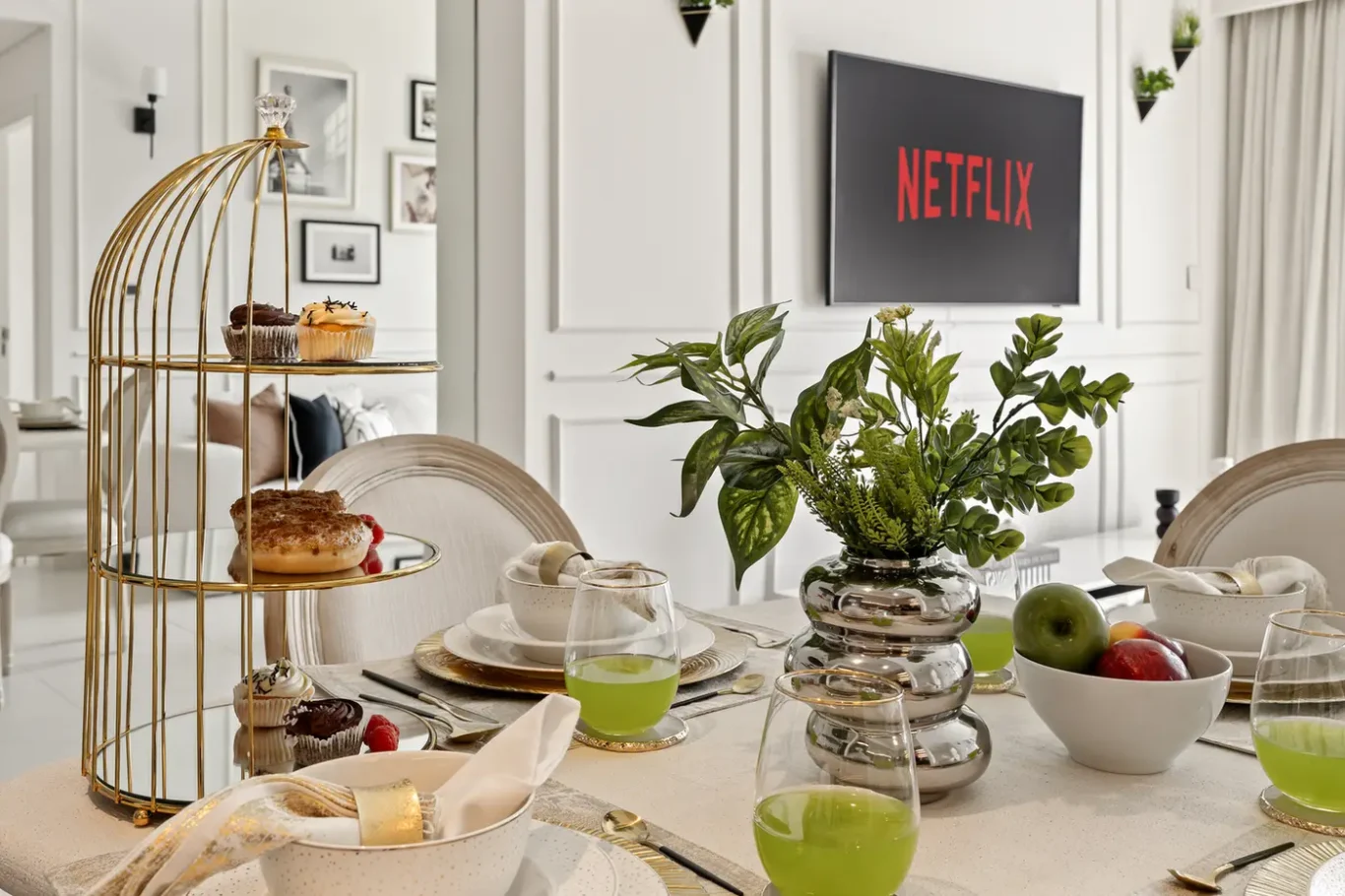 Elegant dining table set for brunch with a gold two-tier pastry stand, green drinks, and a greenery centerpiece, plus a Netflix screen on a white paneled wall.