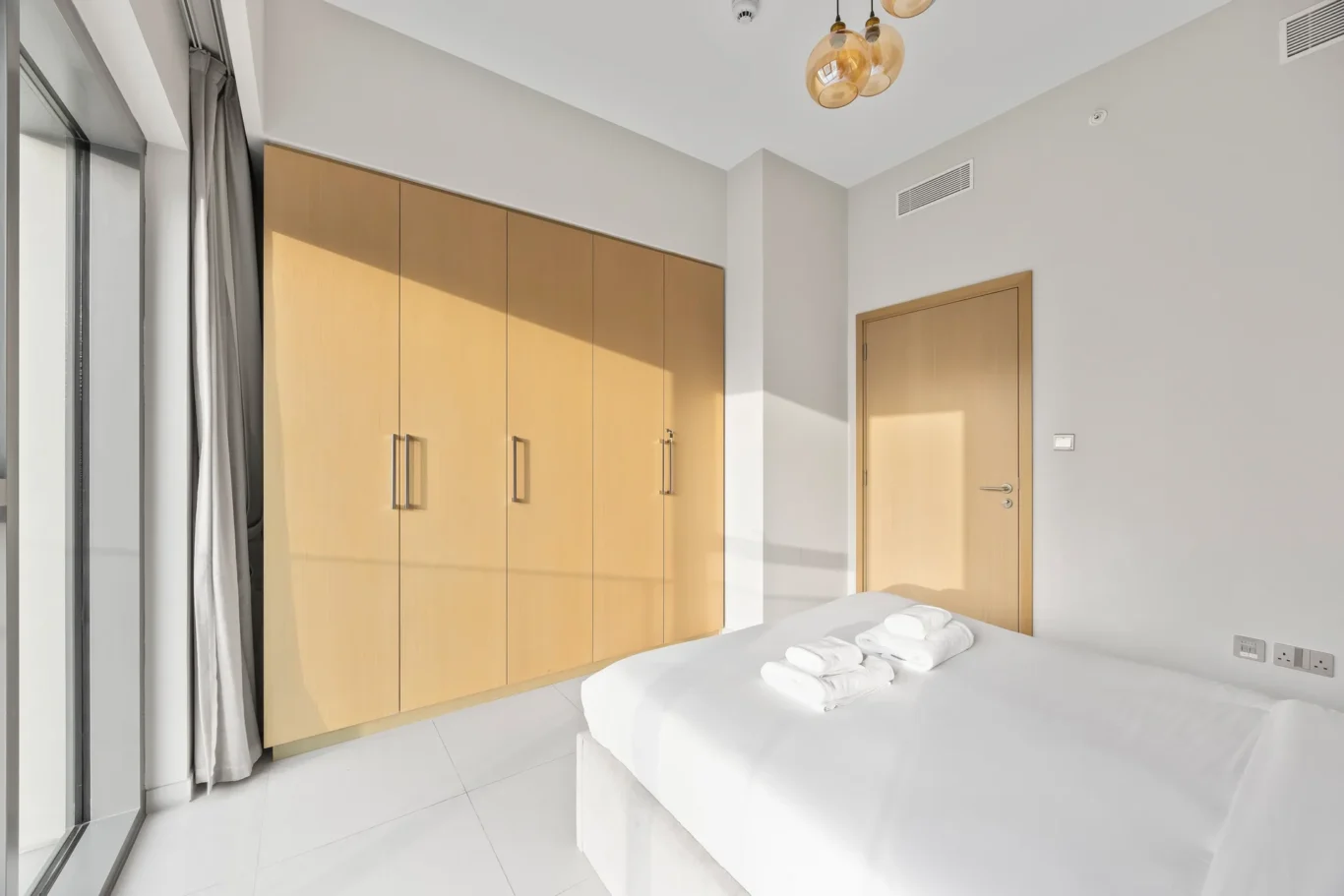 Bedroom with tall beige wardrobe, white bed, folded towels, and sliding glass door to balcony on the left.