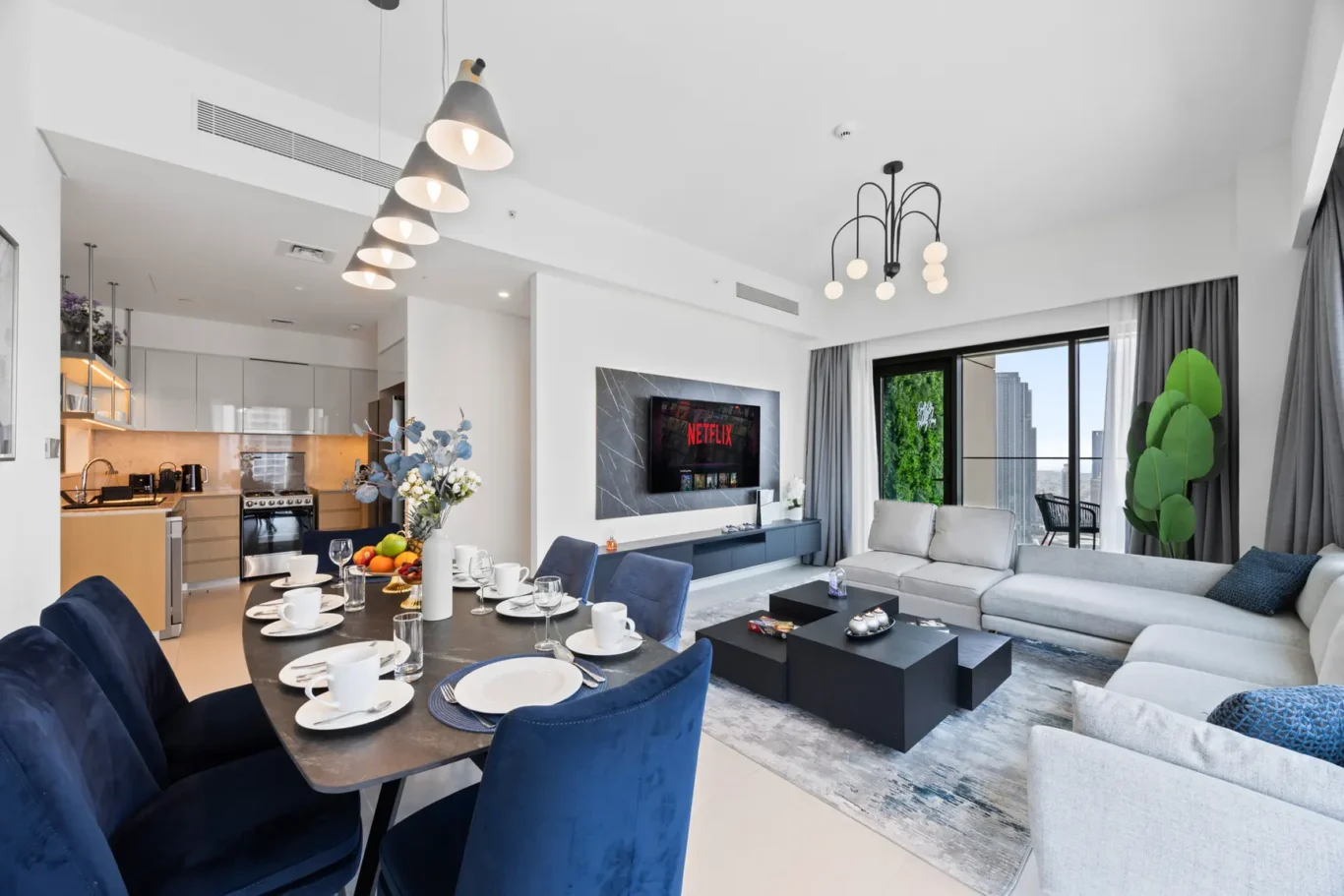 Open-plan living area with a dining table set for six, blue chairs, a gray sofa, and a wall-mounted Netflix screen in a modern kitchen-living space.