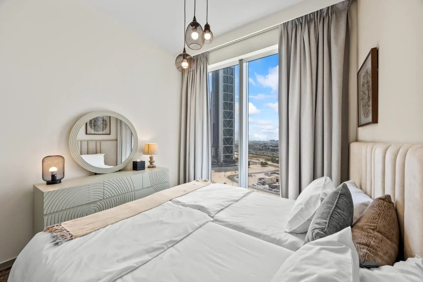 Bright hotel-style bedroom with a large window showing a cityscape, a white dresser with a round mirror, and pendant lights above.