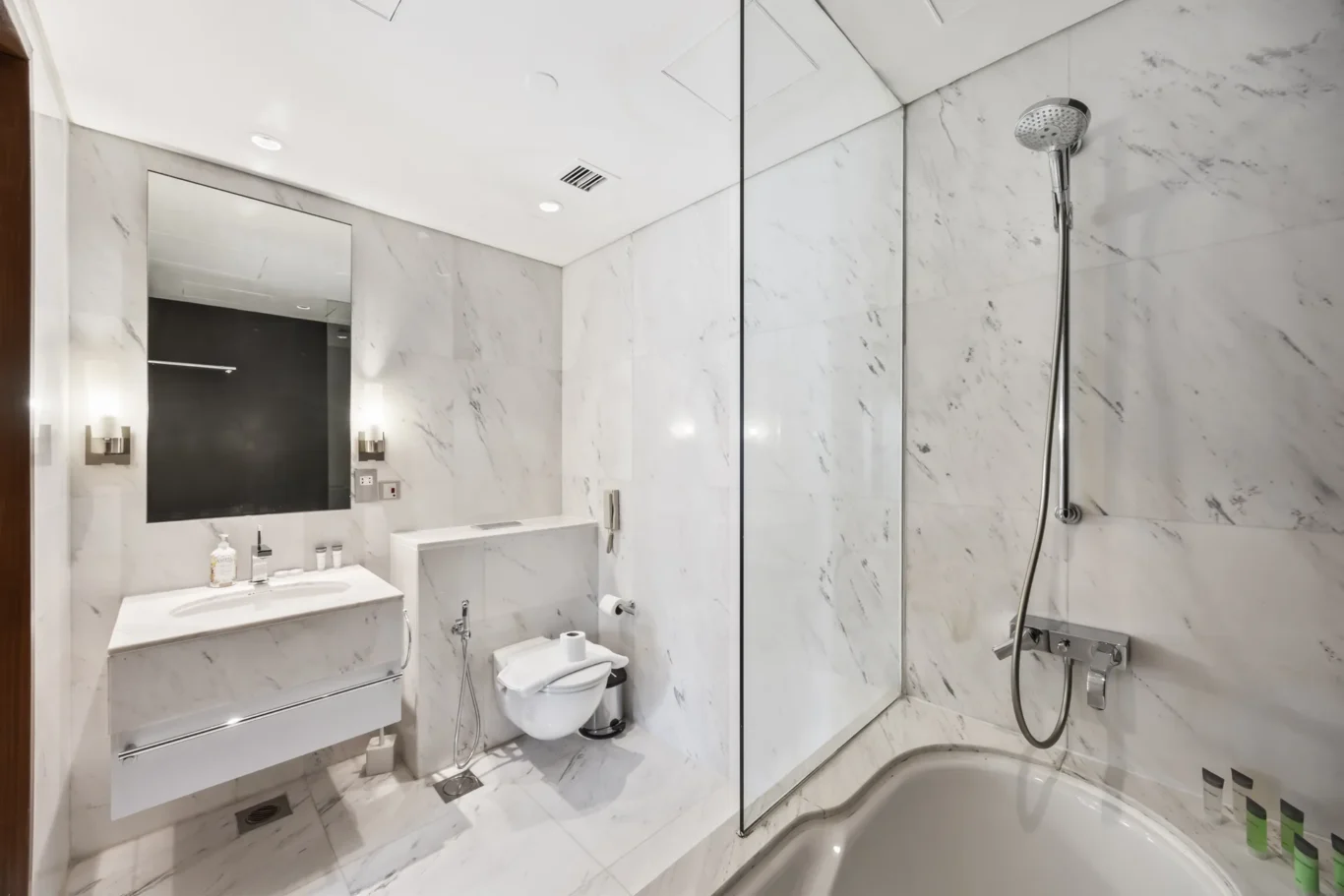 Contemporary marble bathroom with a glass shower enclosure, wall-mounted sink, toilet, and large mirror with chrome fixtures.