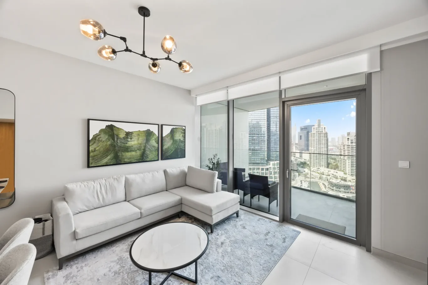 Modern living room with light gray sectional sofa, round marble coffee table, and abstract green wall art near a glass balcony door overlooking a city skyline.