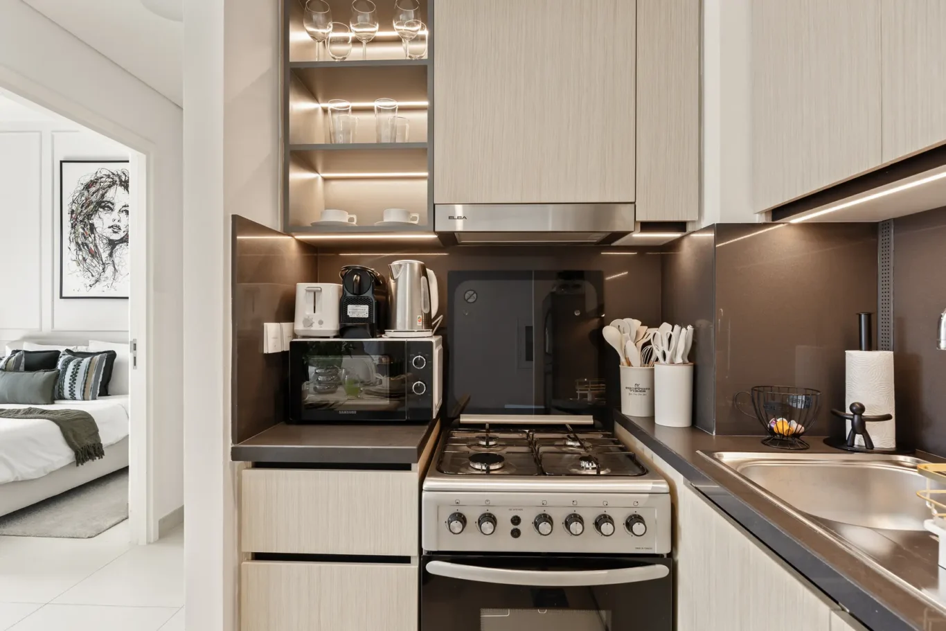 Modern galley kitchen with stainless stove, kettle, coffee machine, and microwave on dark countertops; open shelves with glasses above.
