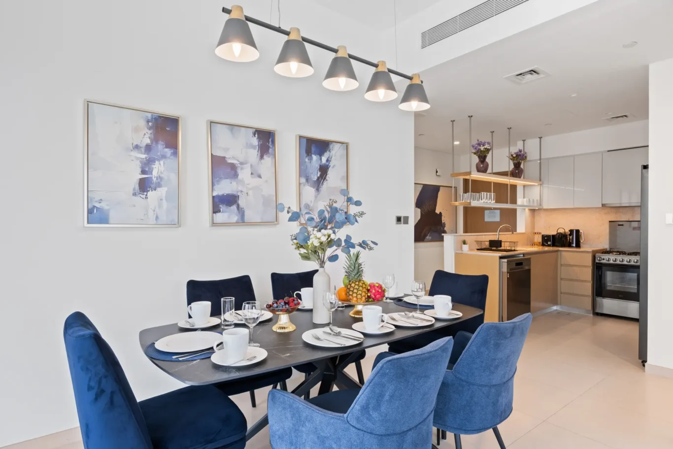 Modern dining room with blue upholstered chairs around a black dining table set for a meal; open kitchen visible in the background with pendant lights above the island.