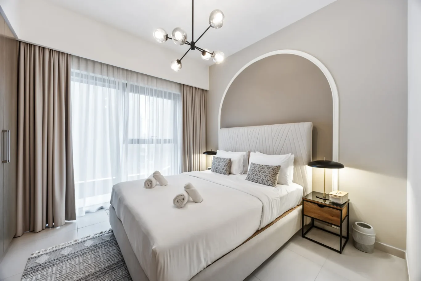 King-size bed with white bedding and decorative pillows beneath a white arched wall niche in a modern hotel-style bedroom.