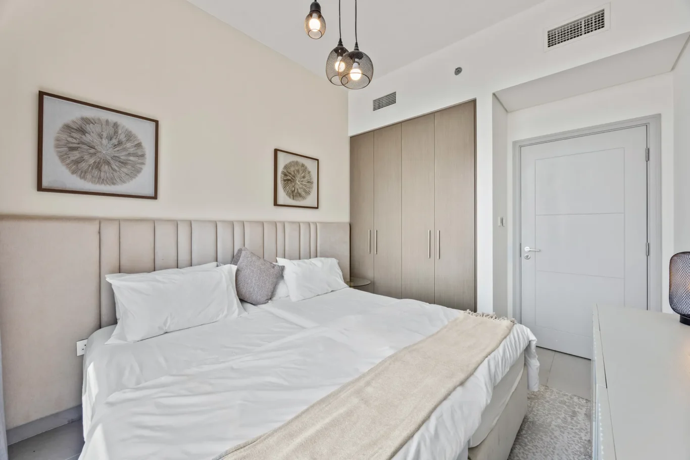 Modern bedroom with a beige upholstered headboard, white bedding, and two framed circular art prints above the bed; built-in wardrobe and a gray door to the right. Consistent neutral tones and pendant lights add style.