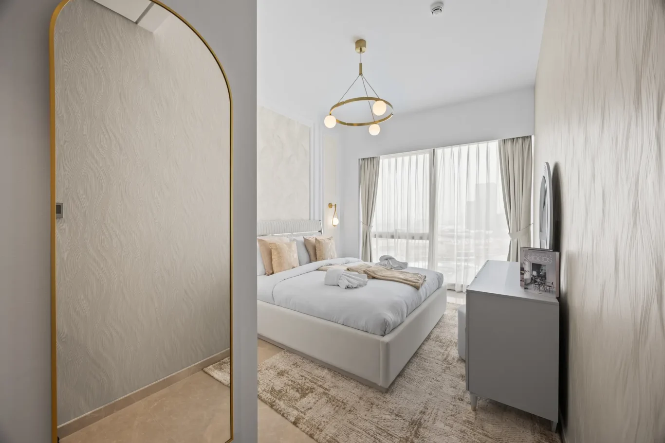 Modern hotel bedroom with a king bed, circular gold chandelier, and gray furniture overlooking large windows with sheer curtains